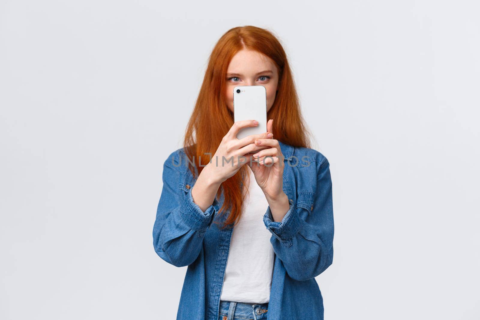 Turning on photo filter. Cheerful and cute female redhead lifestyle blogger, taking picture on smartphone, holding mobile phone near face, taking good shot, searching pretty angle, white background.