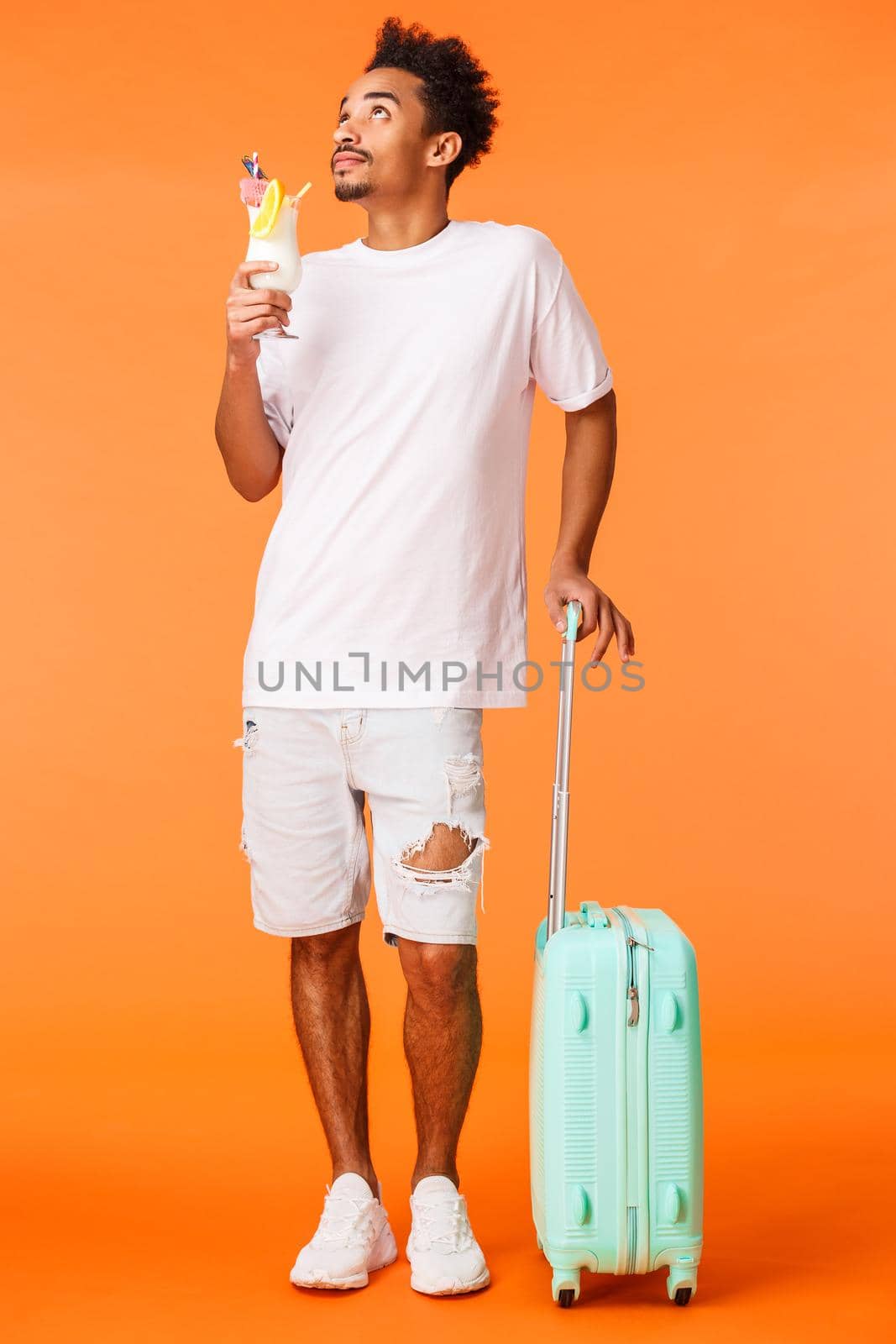 Full-length vertical shot dreamy, relaxed african-american man in white t-shirt, standing near luggage and looking up at sun, drinking cocktail enjoying perfect vacation, standing orange background.