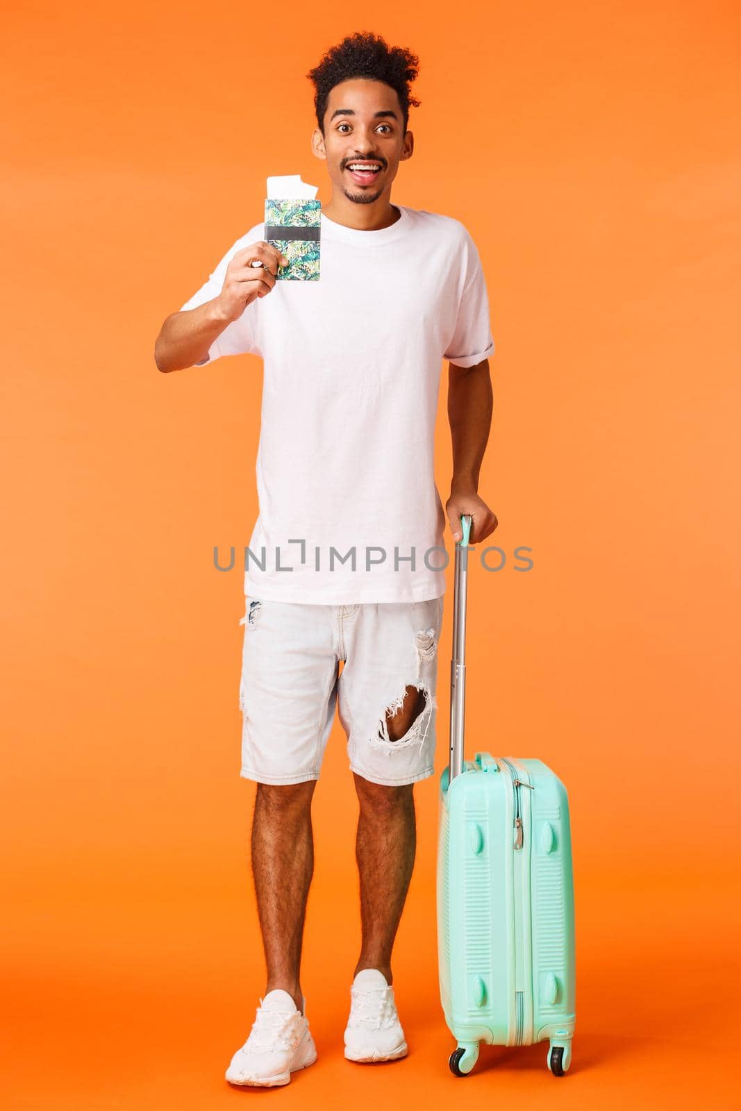 Full-length vertical shot happy upbeat, african american man with luggage, holding passport with two flight tickets, smiling amused, flying abroad, travel on vacation, standing orange background.