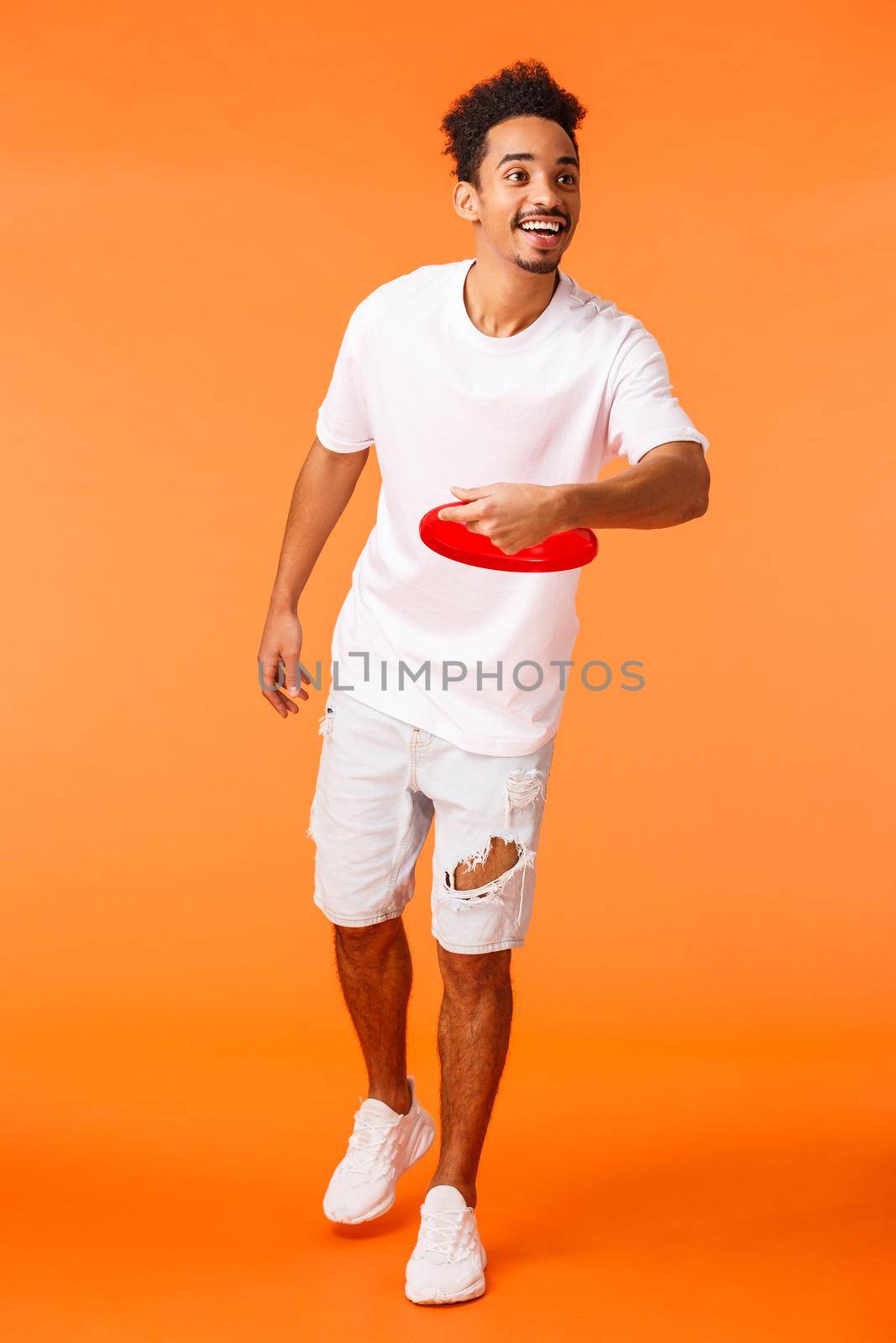 Full-length vertical shot cheerful african-american cute man throwing frisbee and smiling happy, playing game with friends outdoors, enjoying vacation, leisure on holidays, orange background.