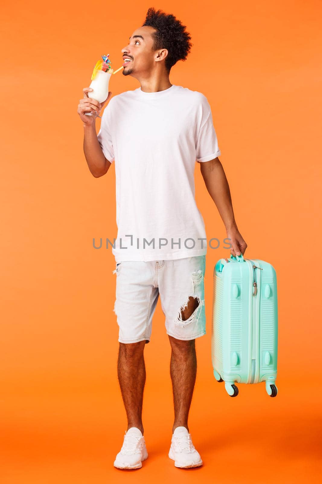 Full-length vertical shot dreamy happy and relaxed african-american man carrying luggage, suitcase and drinking cocktail, looking around pleased and smiling, book in luxury hotel, orange background.