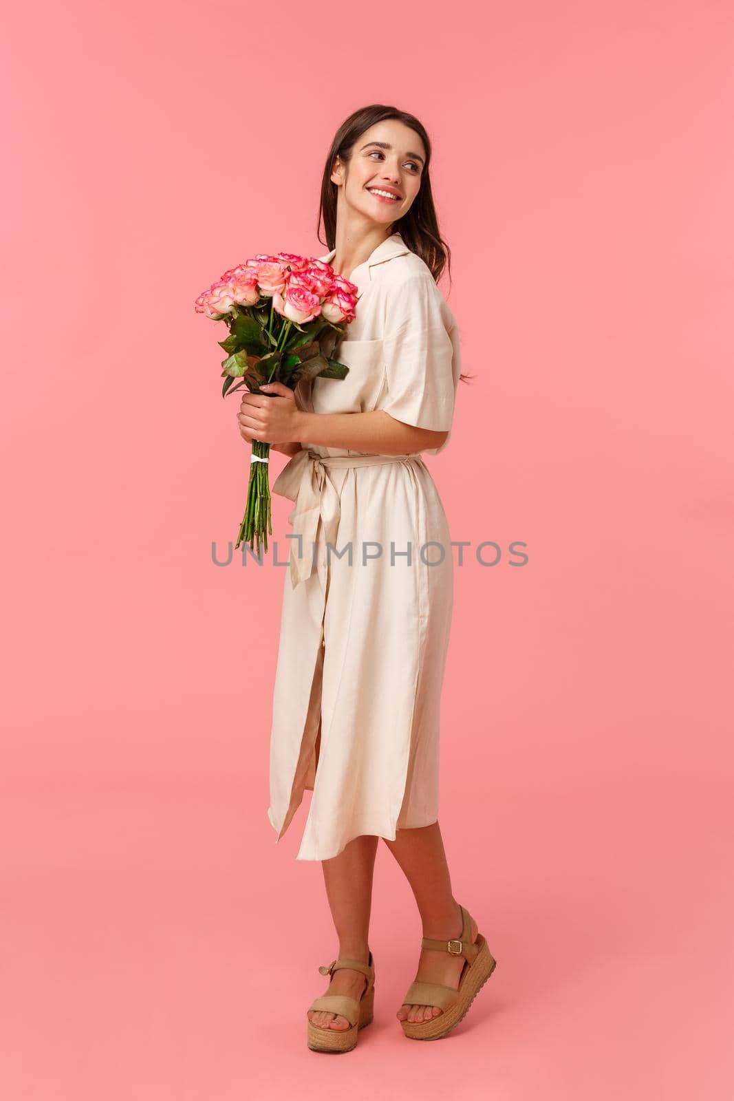 Full-length vertical portrait pretty brunette girl on romantic date receiving nice bouquet flowers, holding roses and gazing behind with delighted soft and tender smile, standing pink background by Benzoix