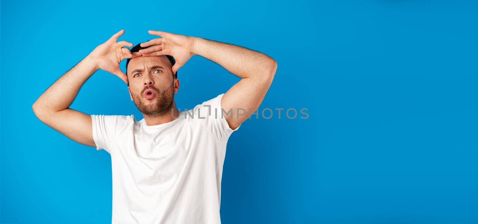 Portrait of a confused young man holding hands on his head over blue background, close up
