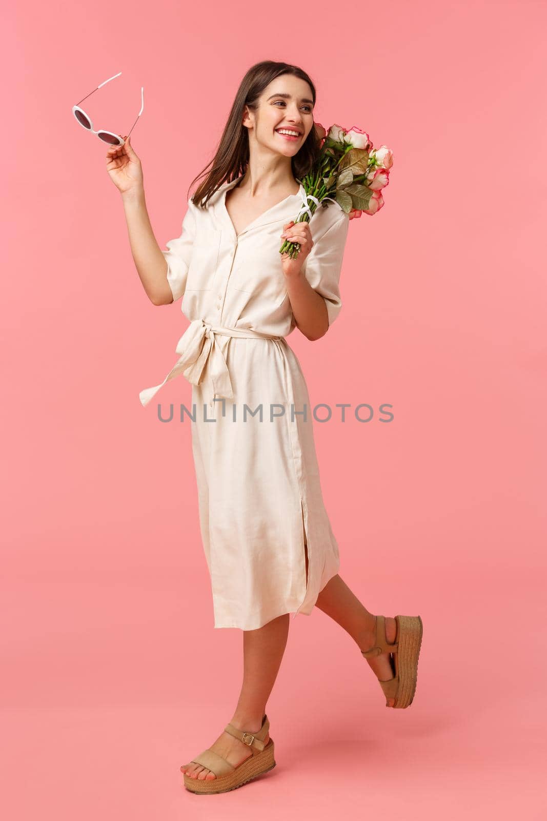 Full-length vertical portrait carefree attractive feminine brunette female in dress, walking along street, receiving beautiful flowers, look behind and smiling, catwalk over pink background.