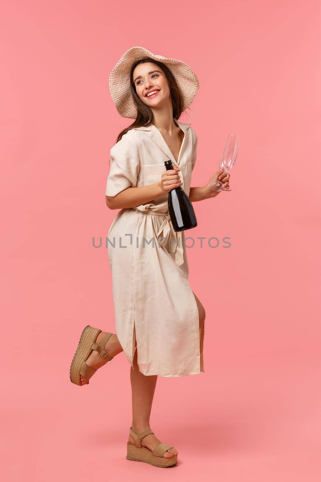 Full-length vertical shot woman on picnic, going out with girlfriend on fine sunny day in hat and dress, rushing with bottle champagne and glasses, smiling happily, standing pink background.