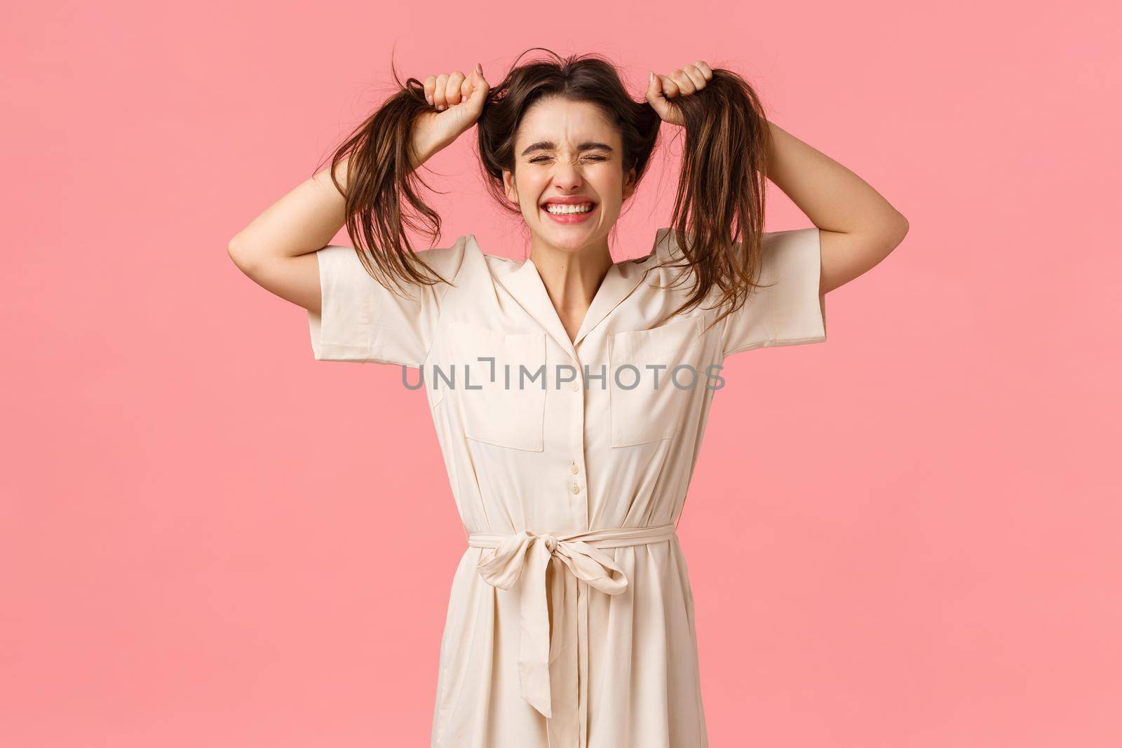 Emotions, celebration and leisure concept. Happy and funny cute girl in dress, messy hair and pulling it, jumping close eyes and smiling joyfully, having fun, fool around playfully, pink background by Benzoix