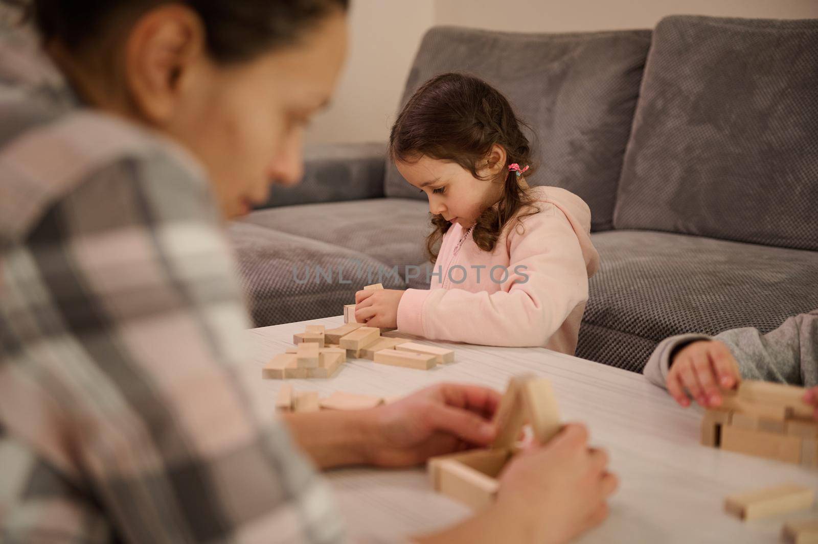 Concentrated little European girl builds constructions with wooden blocks, developing her fine motor skills, sitting at table with her blurred mother on the foreground, focused on building structures