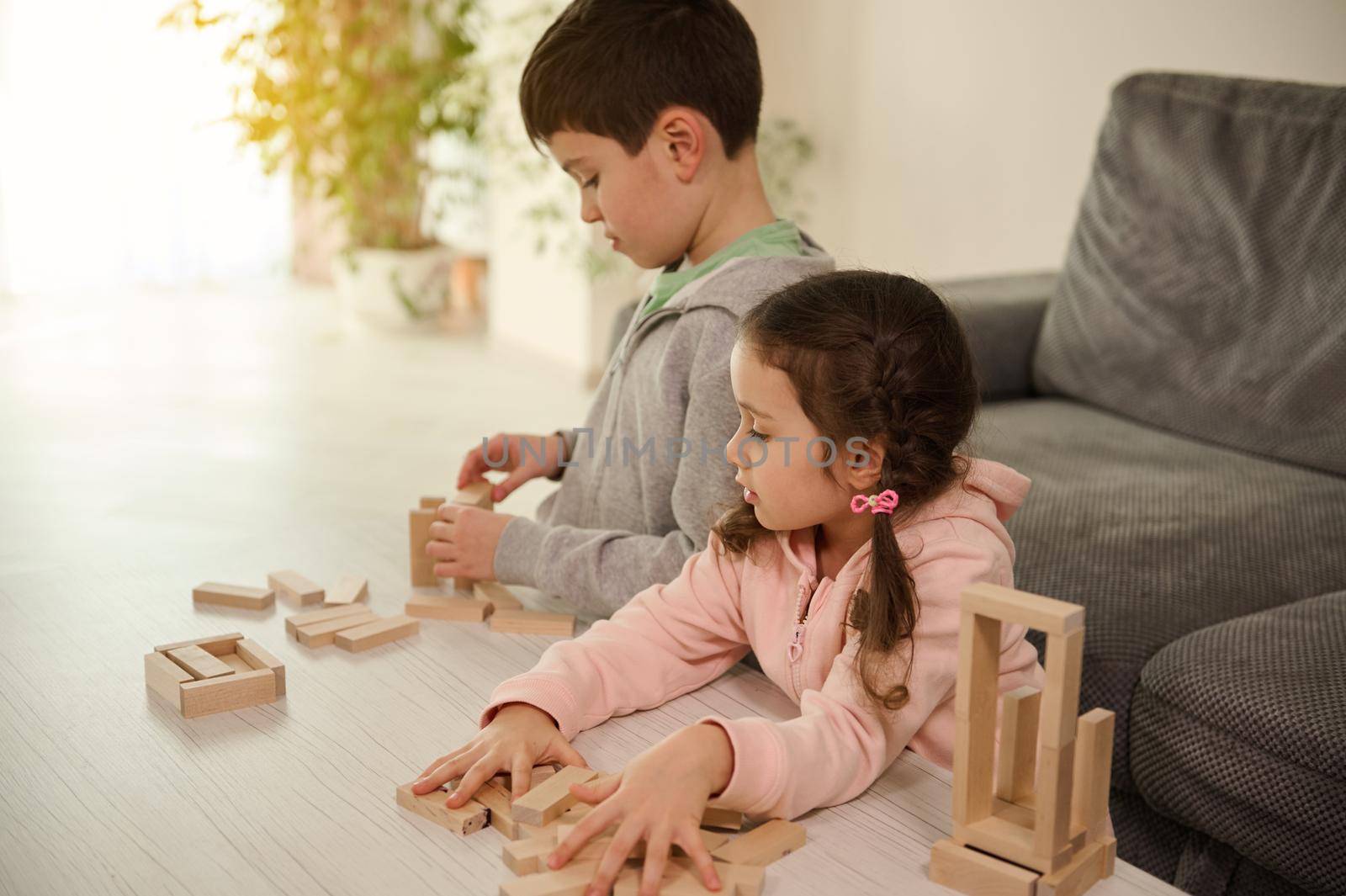 Curious adorable Caucasian girl inspecting her brother's wooden block buildings - childhood activities, fine motors skill development concept. Brother and sister playing board games together at home