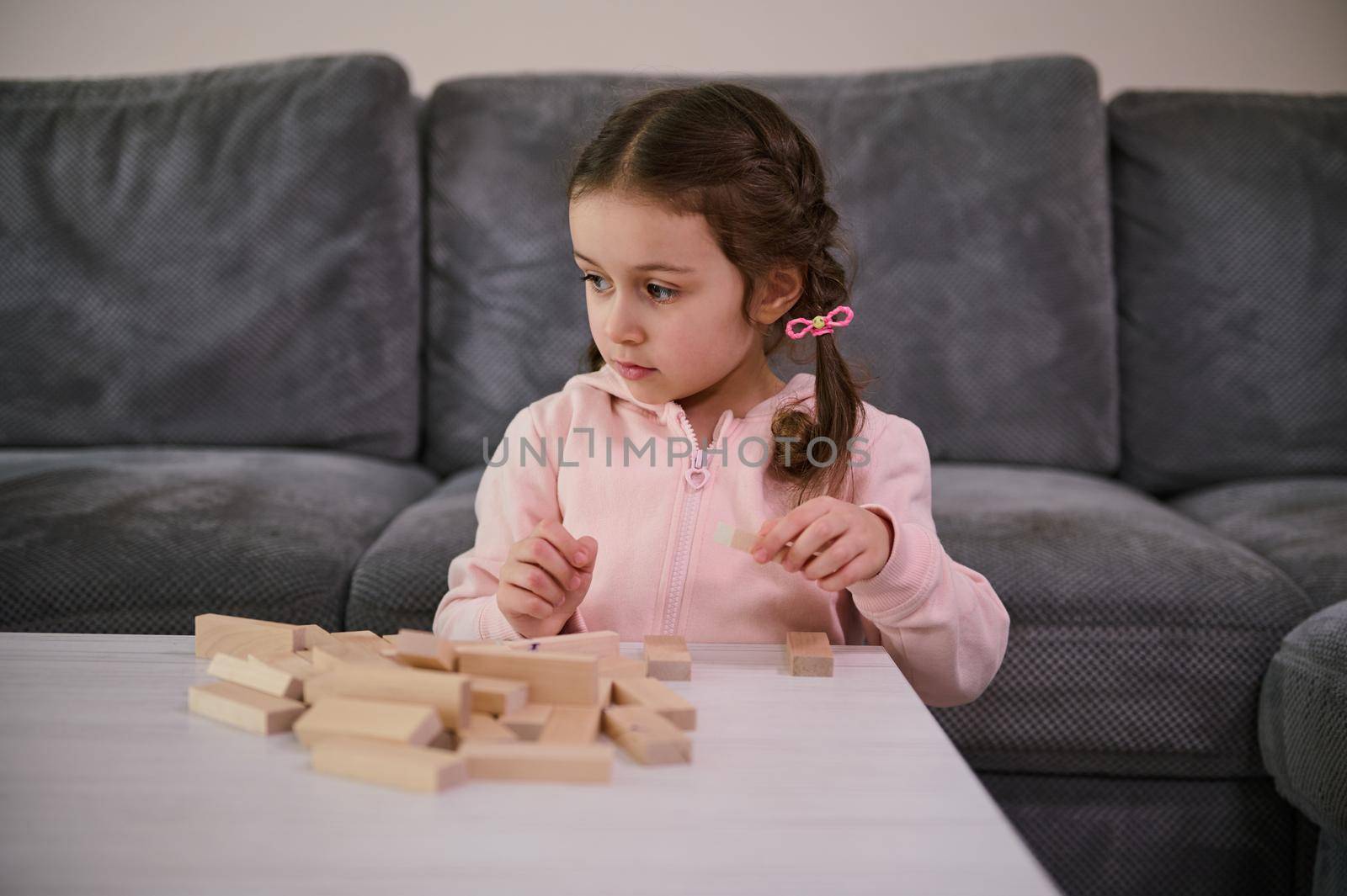 Education, development of fine motor skills, developmental board games concept. Adorable Caucasian girl focused on building with wooden bricks blocks, sitting at a table in the home living room