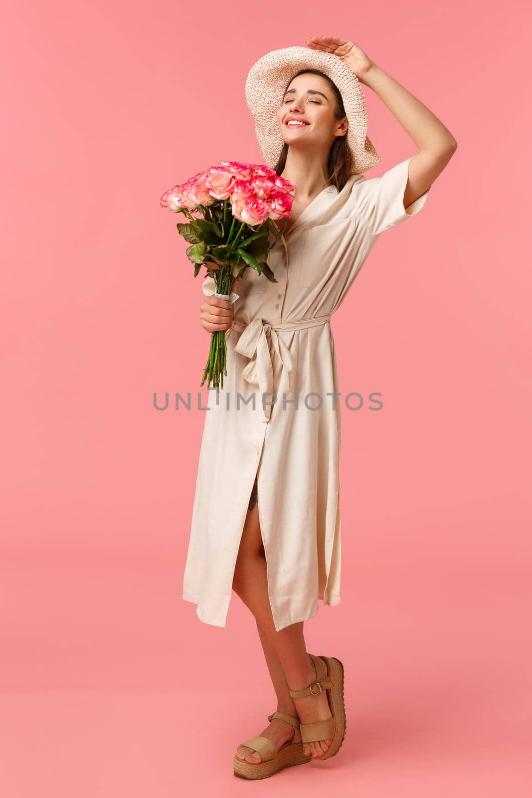 Full-length vertical portrait charming, romantic woman in dress and hat, holding flowers and close eyes as leaning towards sun, enjoying beautiful warm day, having perfect weekend, pink background.