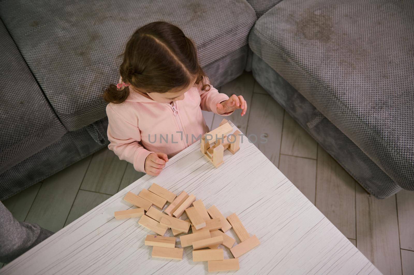 Overhead view of creative preschool child, concentrated baby girl on building with wooden blocks bricks. Educational board game and developmental smart pastime. Fine motor skill development concept