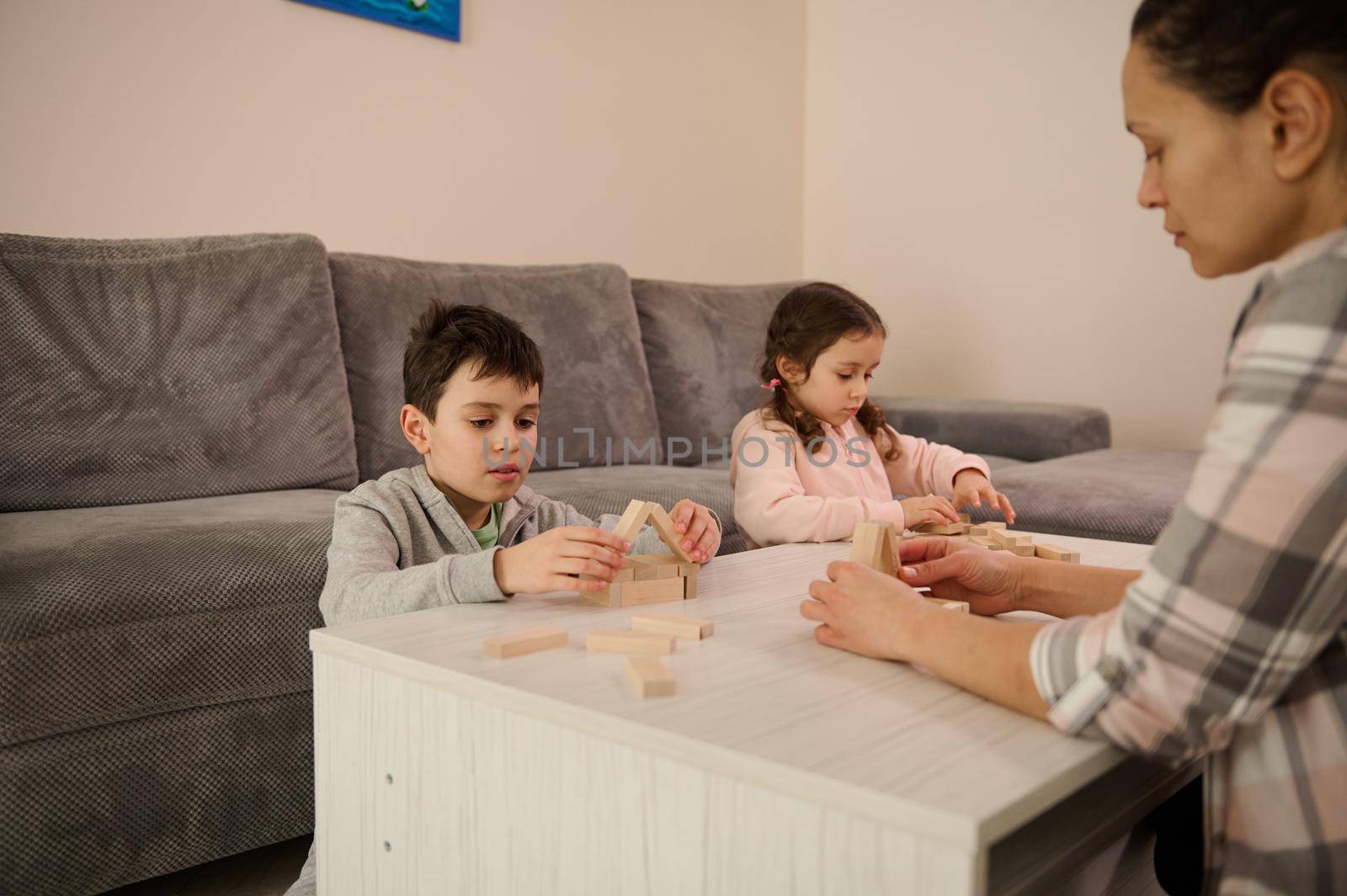 Focus on a handsome school age boy building wooden house with blocks and bricks, sitting at table near her sister and mom focused on construction of structures. Family pastime and leisure concept by artgf