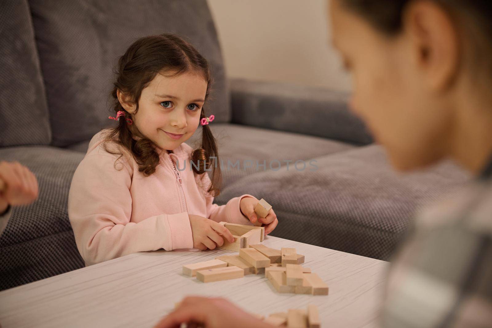 Cute baby girl in pink sweatshirt playing board game, building wooden structures with blocks, smiles looking at her mom. Family pastime, educational leisure and fine motor skills development concept by artgf