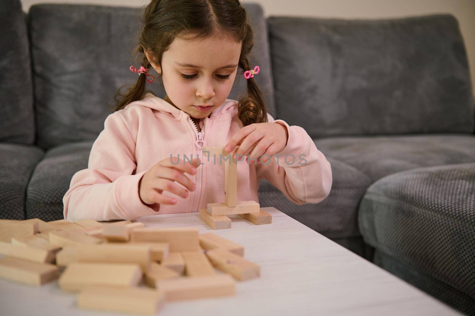 Adorable little girl with two pigtails, dressed in pink sweatshirt playing board game, building wooden constructions with blocks. Hand movement control and building computational skills concept.