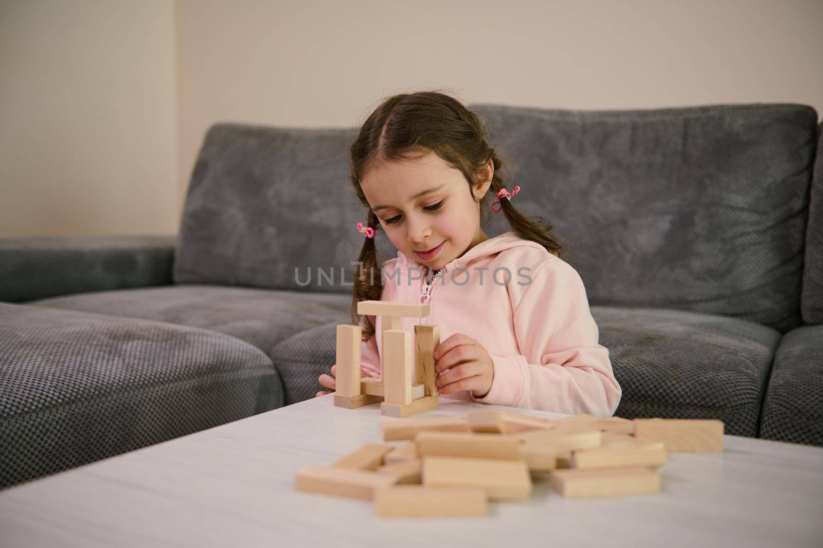Cheerful creative beautiful European 5 y.o. little girl with two pigtails in pink sweatshirt playing educational developmental board game, she's concentrated on building wooden structures with blocks