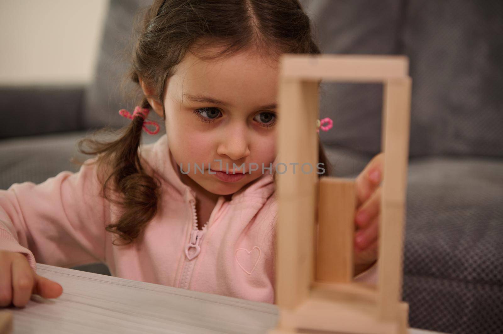 Close-up portrait of adorable Caucasian child, preschool girl in pink sweatshirt concentrated on building wooden structure with wooden blocks. Board games and fine motor skills development concept