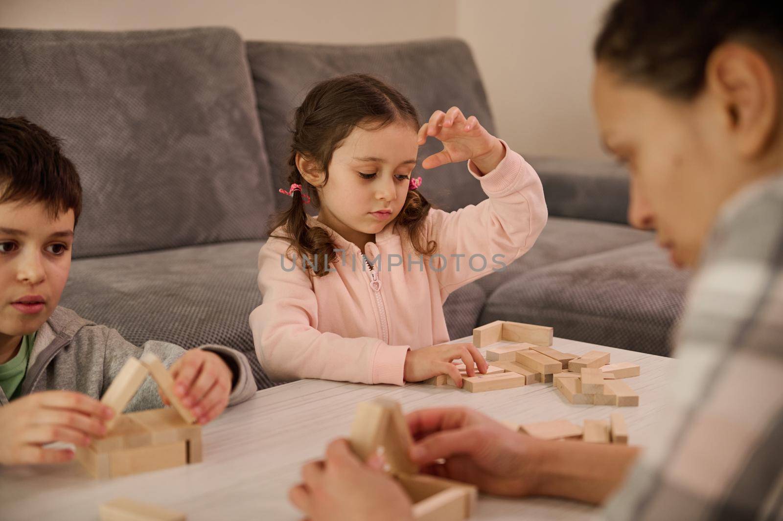 Fine motor skills, concentration development, educational leisure, family pastime concept. Cute kids playing board game with their blurred mom focused on building a wooden structure in the foreground by artgf