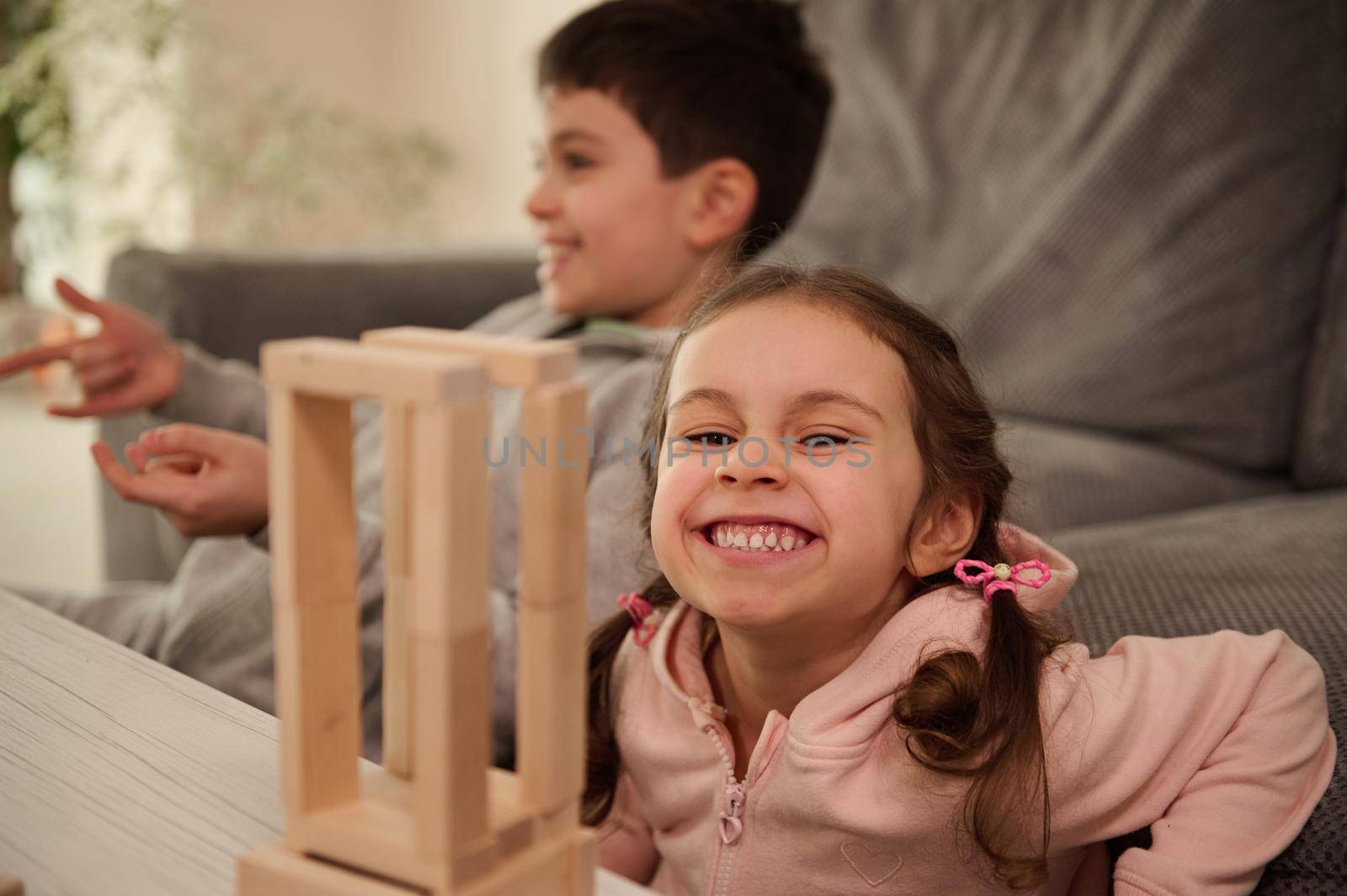Adorable child, beautiful little girl in pink sweatshirt plays board game with her brother, builds wooden structures from wooden bricks and blocks, smiles toothy smile, looking at camera
