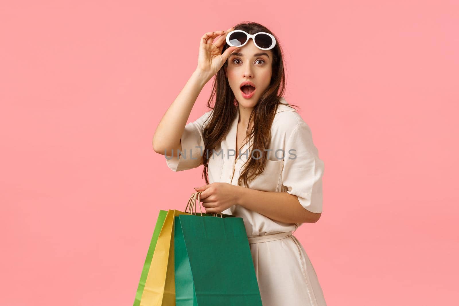 Amused feminine girl, waisting all money on new clothes, found favorite store, holding shopping bags and gazing camera excited, taking-off glasses seeing something fantastic, pink background.