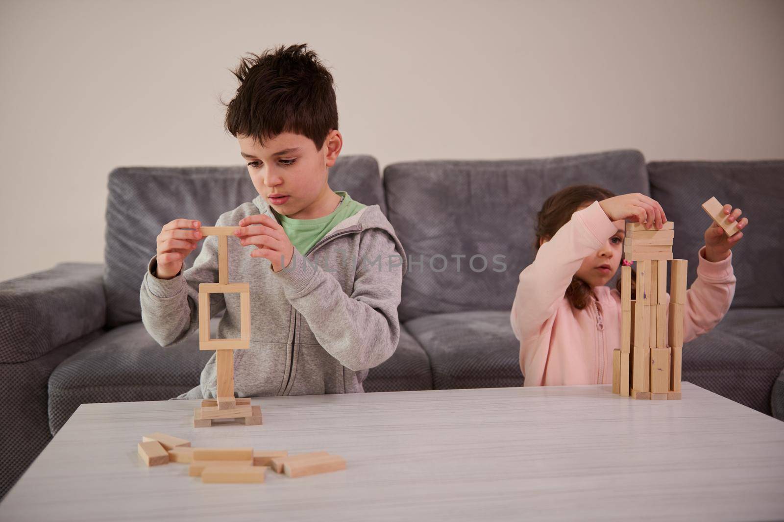 Creative children, brother and sister concentrated on building from wooden blocks, playing developmental board games, building complex tall wooden structures at home. Educational leisure and pastime by artgf