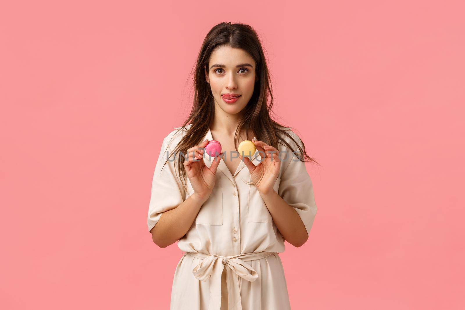 Holidays, beauty and sweets concept. Charming caucasian woman in dress, holding macarons and licking lips from desire try dessert, visit favorite candy store, standing pink background.