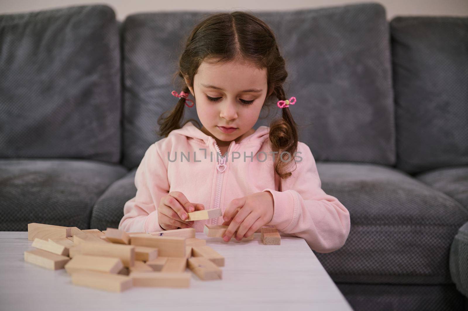 Education, development of fine motor skills, developmental board games concept. Adorable Caucasian girl focused on building with wooden bricks blocks, sitting at a table in the home living room by artgf