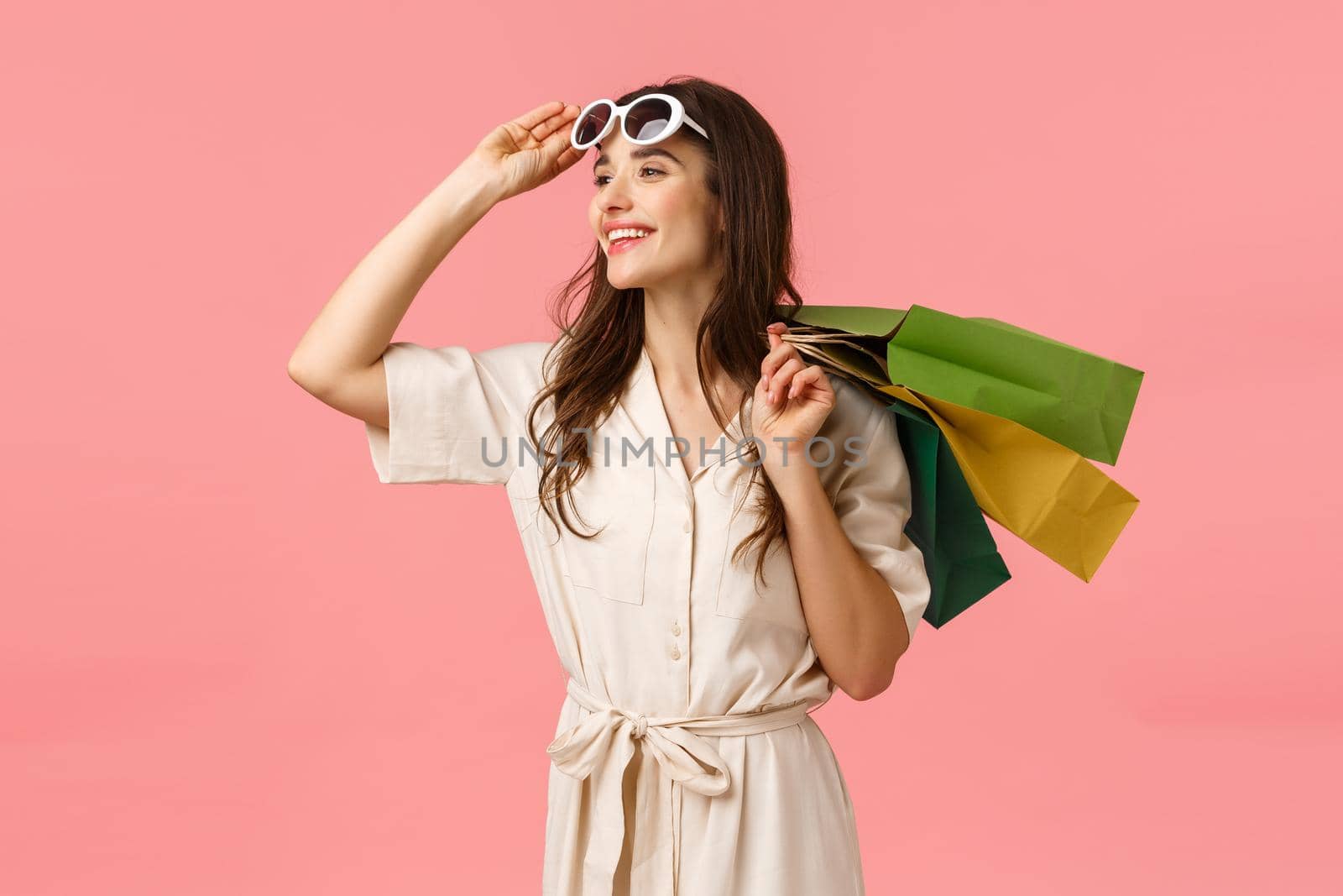 Carefree enthusiastic woman in dress, taking off glasses and looking happily sideways as enjoying beautiful day, fantastic shopping with discounts, holding shop bags behind back, pink background.