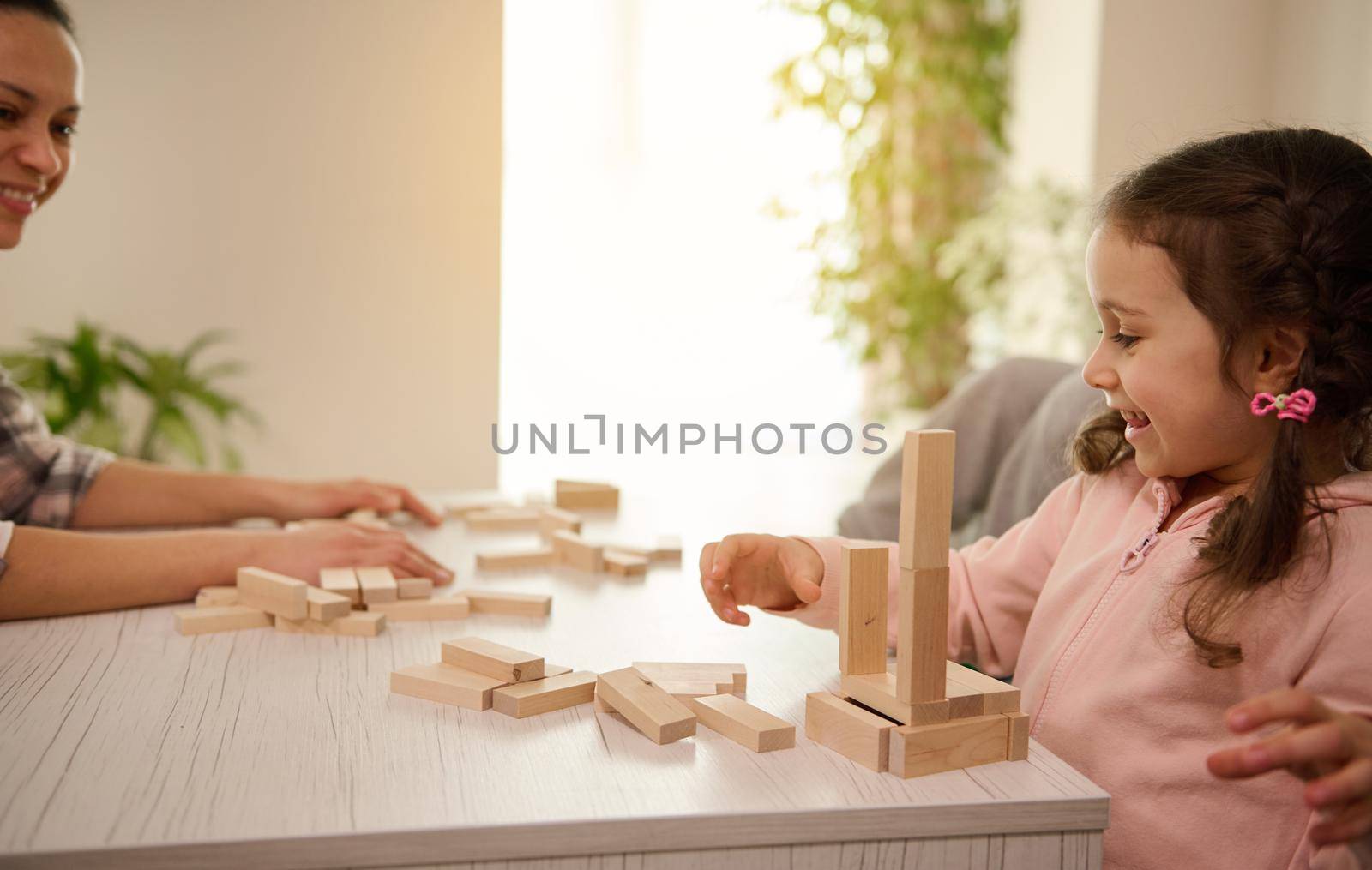 Beautiful child, adorable little girl in pink sweatshirt plays board game with her mother, builds wooden structures from wooden bricks and blocks, smiles toothy smile, enjoying her educational pastime by artgf
