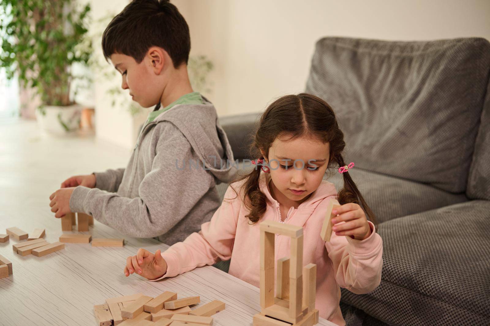 Two adorable Caucasian kids, concentrated children, boy and girl playing board games, building wooden constructions on the table at home. Educational leisure and family pastime concept
