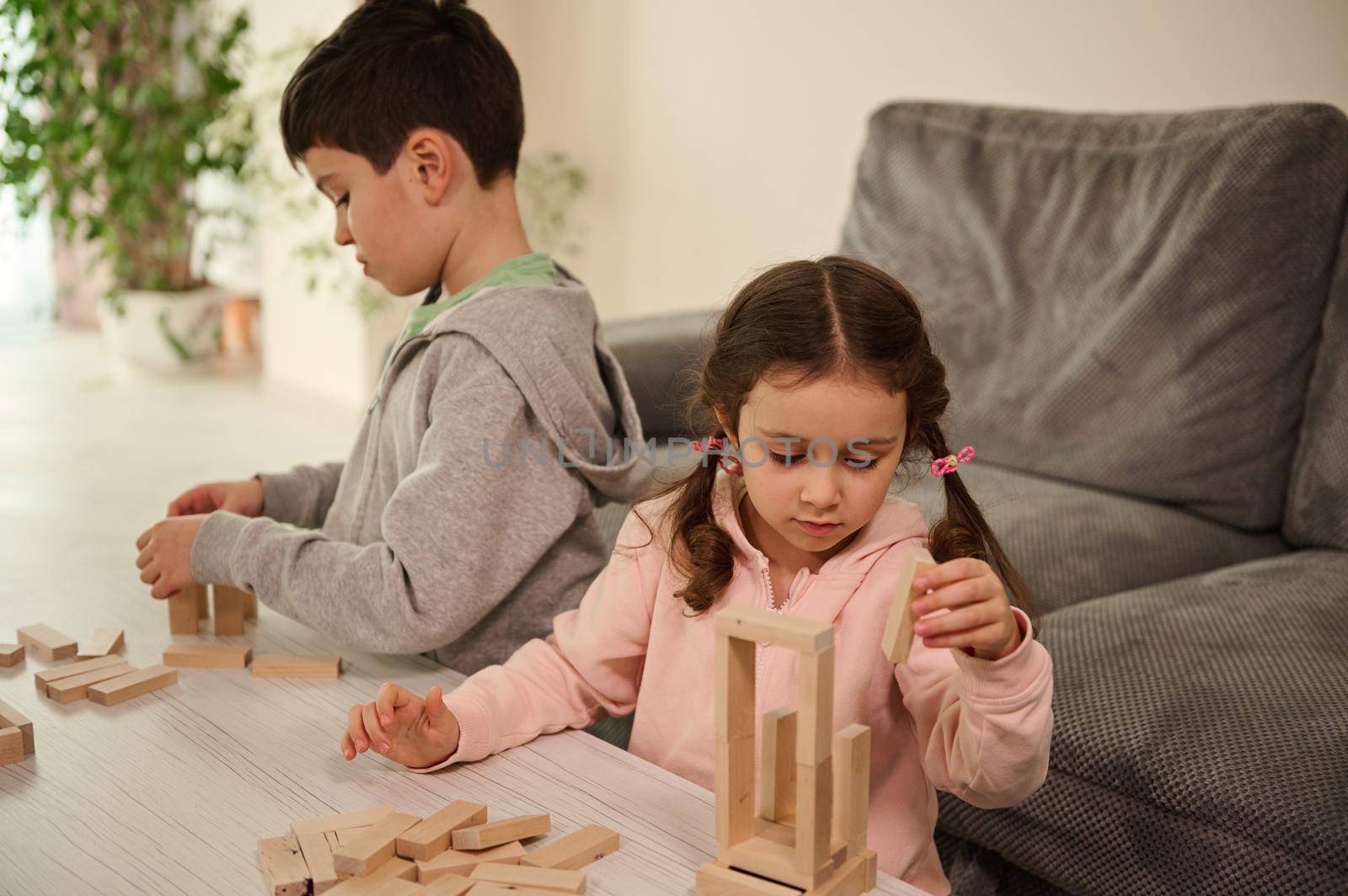 Two adorable Caucasian children, concentrated kids, boy and girl playing board games, building wooden constructions on the table at home. Educational leisure and family pastime concept by artgf