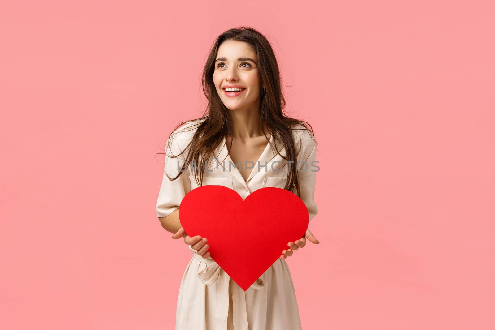 Amused and carefree good-looking european female in dress, holding valentines card, big red heart, looking left amazed and happy, smiling impressed, standing pink background joyful.