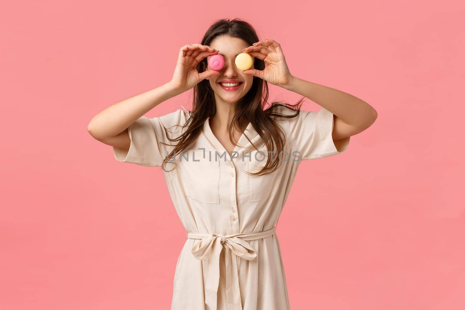 Positivity, delicious food and emotions concept. Happy and carefree european girl like eating sweets, making eyes from macarons and smiling upbeat, standing pink background joyful.