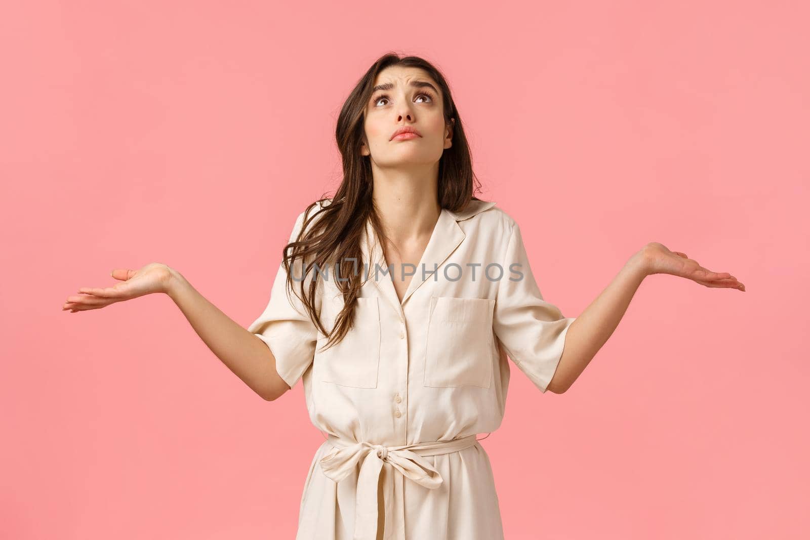 Girl cant understand what faith prepared. Confused and indecisive glamour young brunette girl in dress, shrugging with hands sideways, looking up pouting rain, pink background by Benzoix