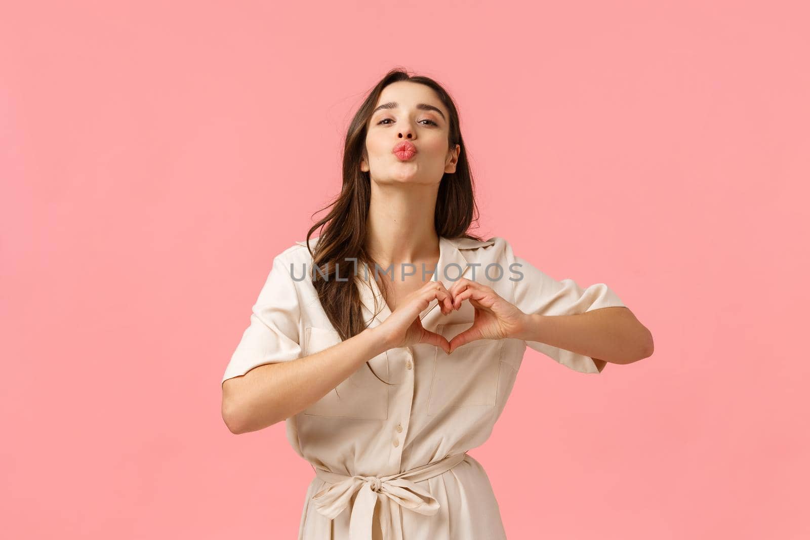 Fashion, beauty and tenderness concept. Charming gorgeous brunette female in cute dress, showing heart love gesture, folding lips for kiss, express romantic feelings, affection, pink background.