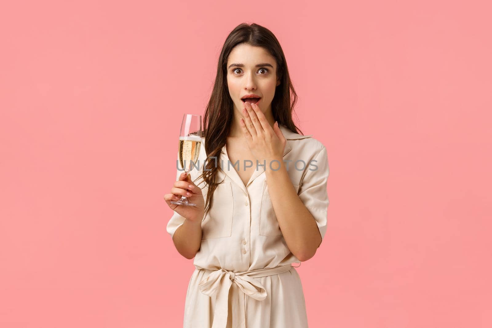 Girl gossiping on party with girlfriends. Attractive amused and surprised young lady in dress, holding champagne glass, open mouth wondered cover with hand and staring camera astonished.