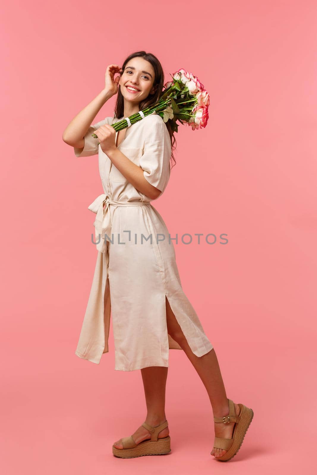 Full-length vertical portrait, dreamy romantic brunette female enjoying date, holding bouquet roses, touching hair tenderly, smiling silly camera, receiving flowers, standing pink background.