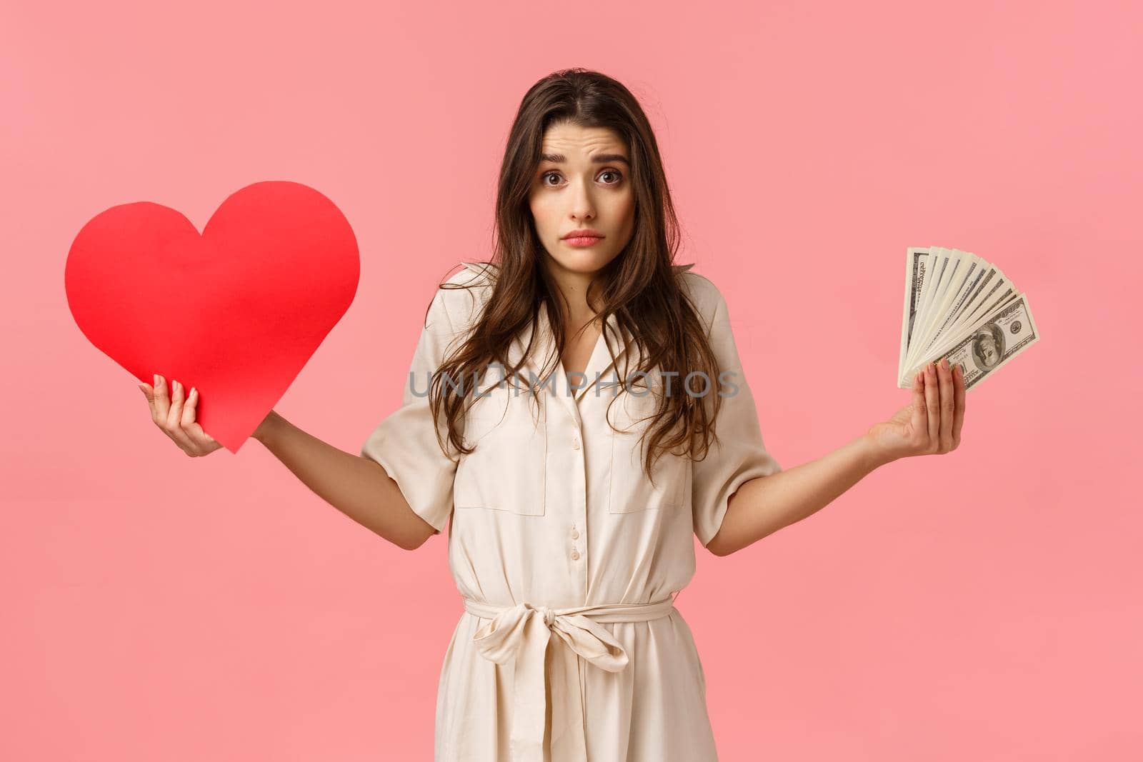 Indecisive and unsure, cute lovely modern woman cant decide what important, shrugging looking confused camera, holding heart card and money, dont know whats right, questioned over pink background.