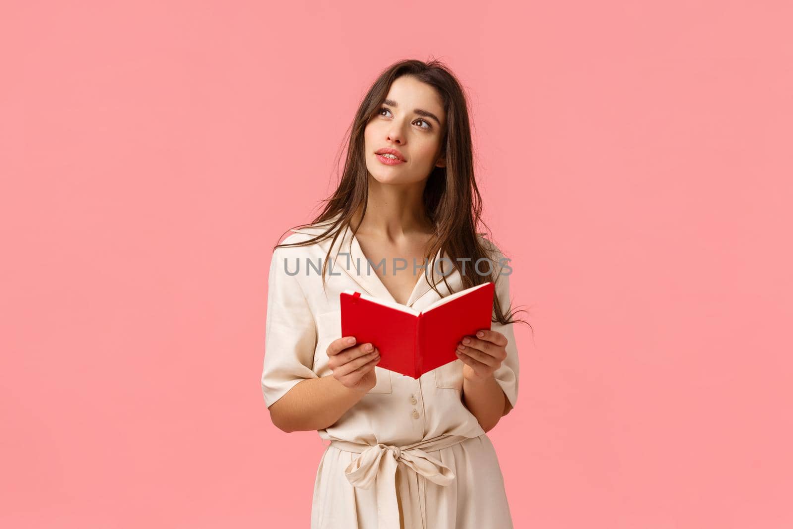 Thoughtful and inspired, creative female student having inspiration, looking up dreamy and wondered, holding red notebook, learning or preparing to class, standing pink background.