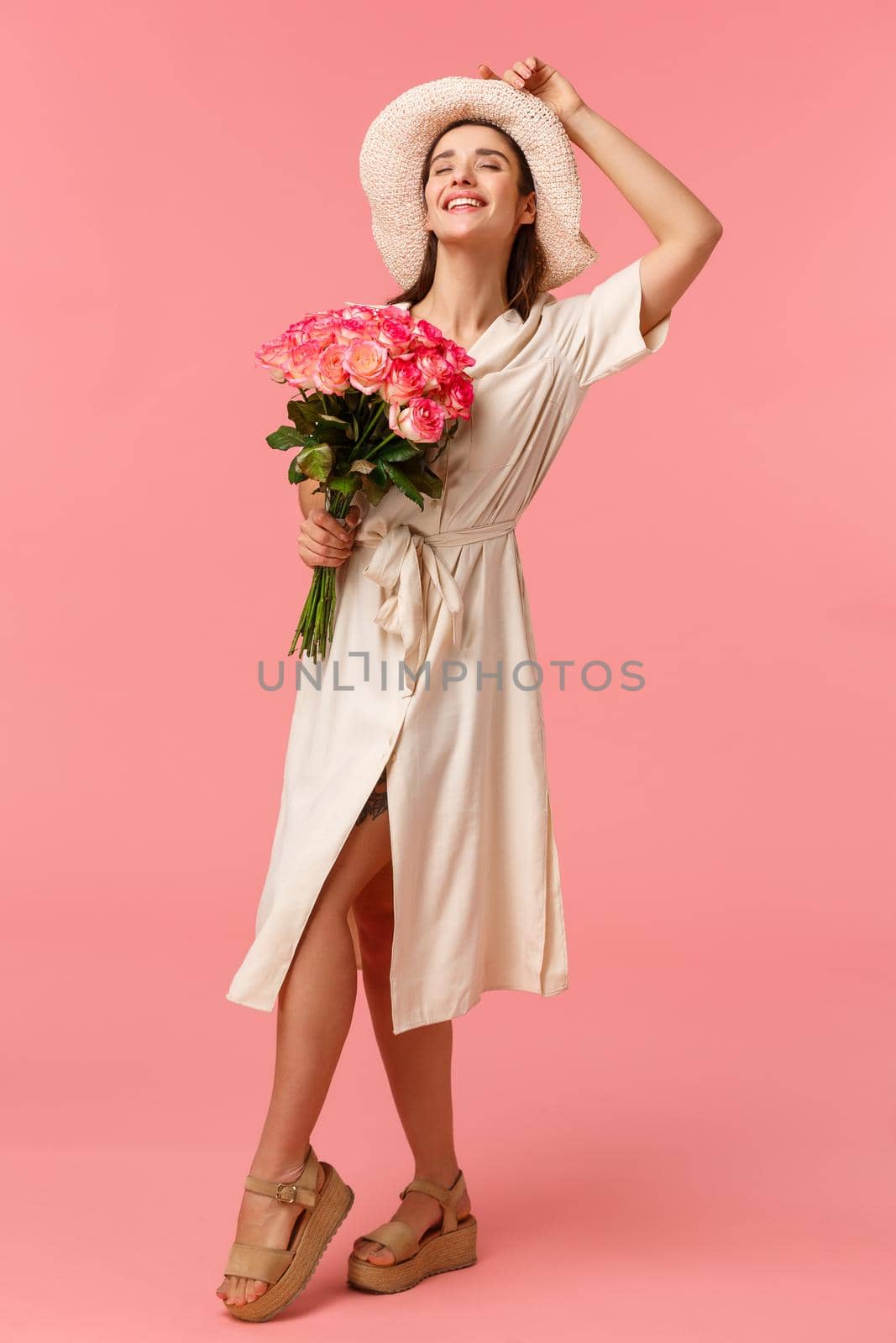 Full-length vertical portrait gorgeous stylish young european woman in dress and hat, close eyes and smiling as enjoying warm sunlights on her face, holding bouquet roses, pink background by Benzoix