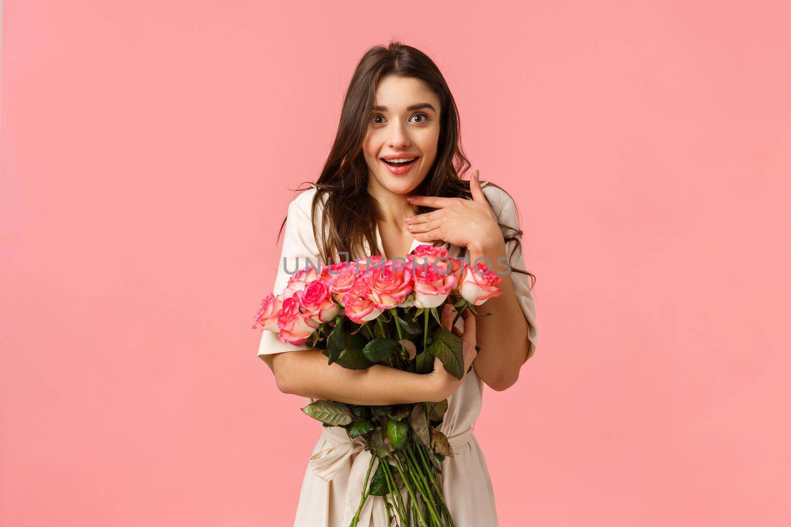 Surprised girl astonished and touched with beautiful roses boyfriend brought on date. Attractive young woman in dress, smiling and gasping flattered, holding stunning bouquet, pink background.