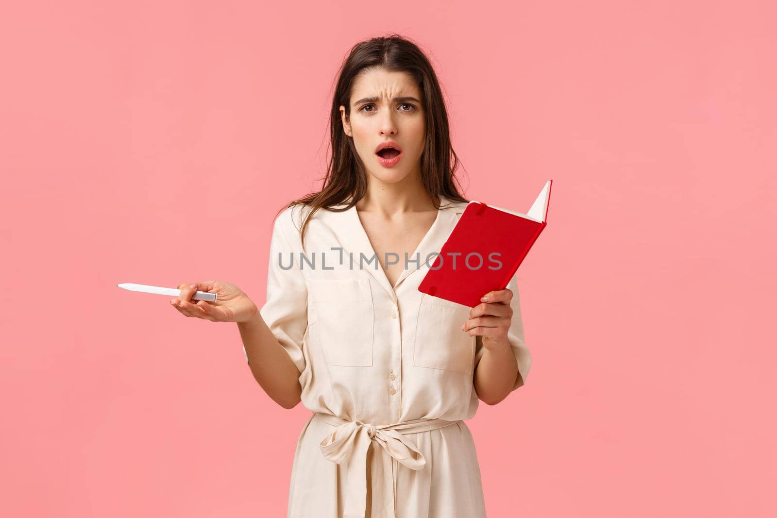Confused and troubled unsure brunette female looking at camera frustrated and displeased, reading someones diary, holding red notebook and pen, complaining, standing pink background.