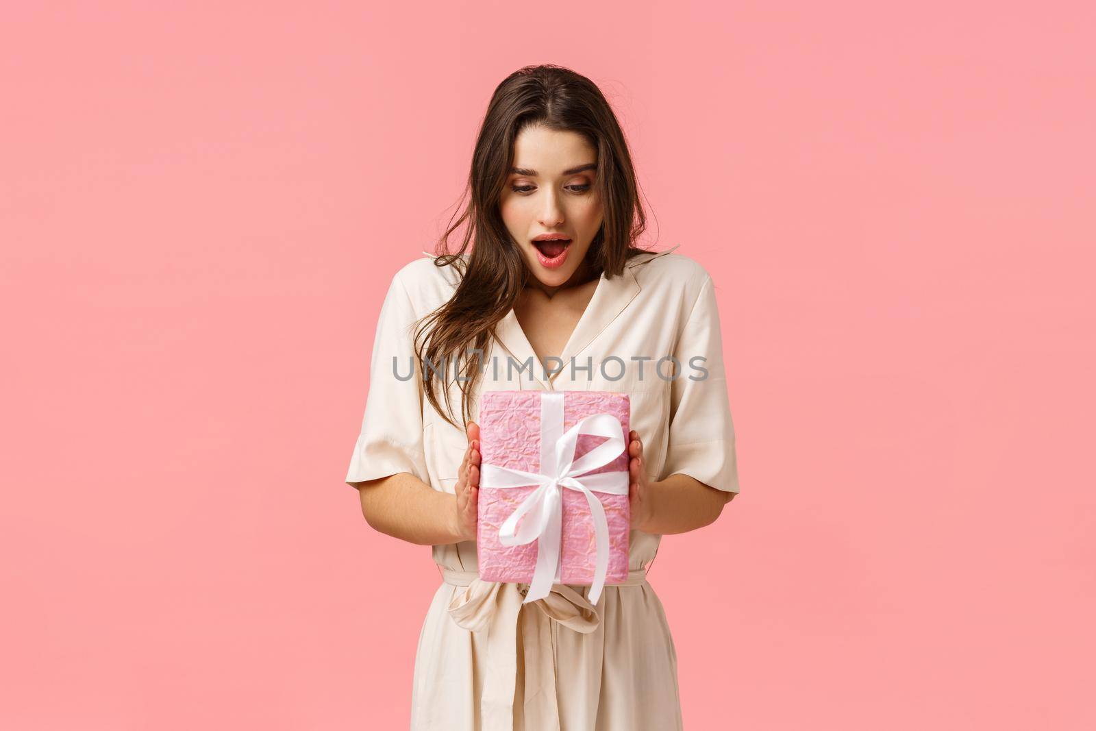Amazement, celebration and holidays concept. Surprised girlfriend holding box of present, didnt expact receive gift, open mouth fascinated look touched wrapped box, pink background by Benzoix