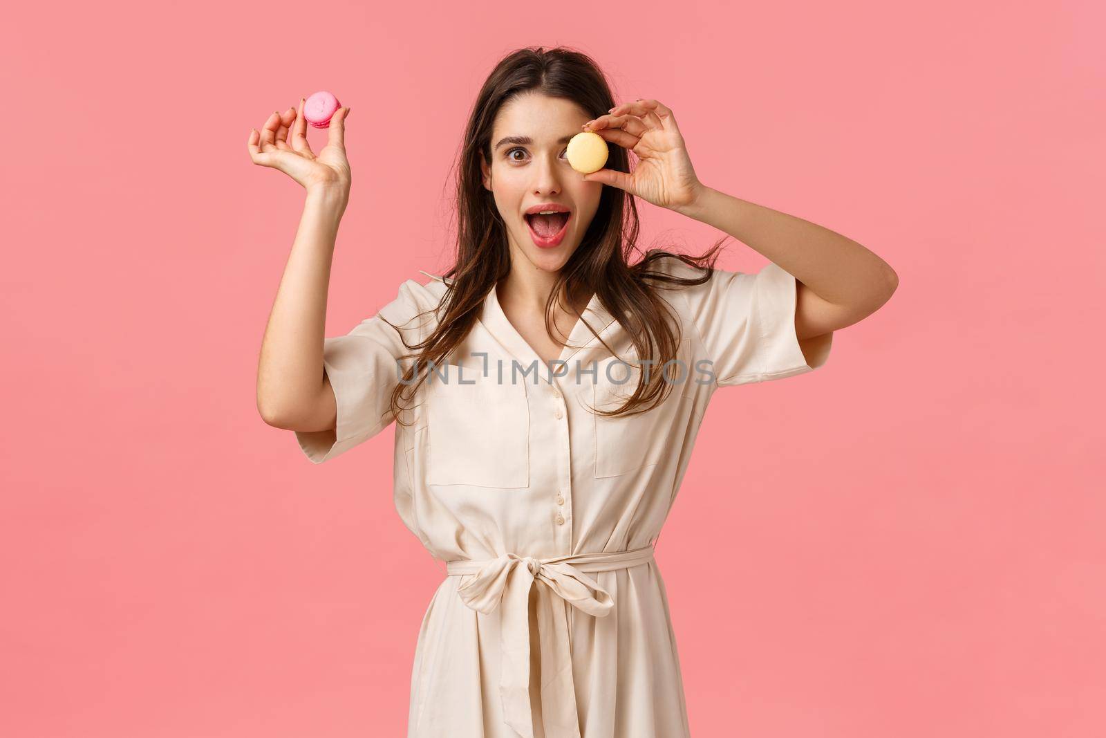 Excitement, holidays and sweet desserts concept. Elegant attractive young woman enjoy eating delicious food, holding macarons on on eye, open mouth gasping amused, standing pink background.