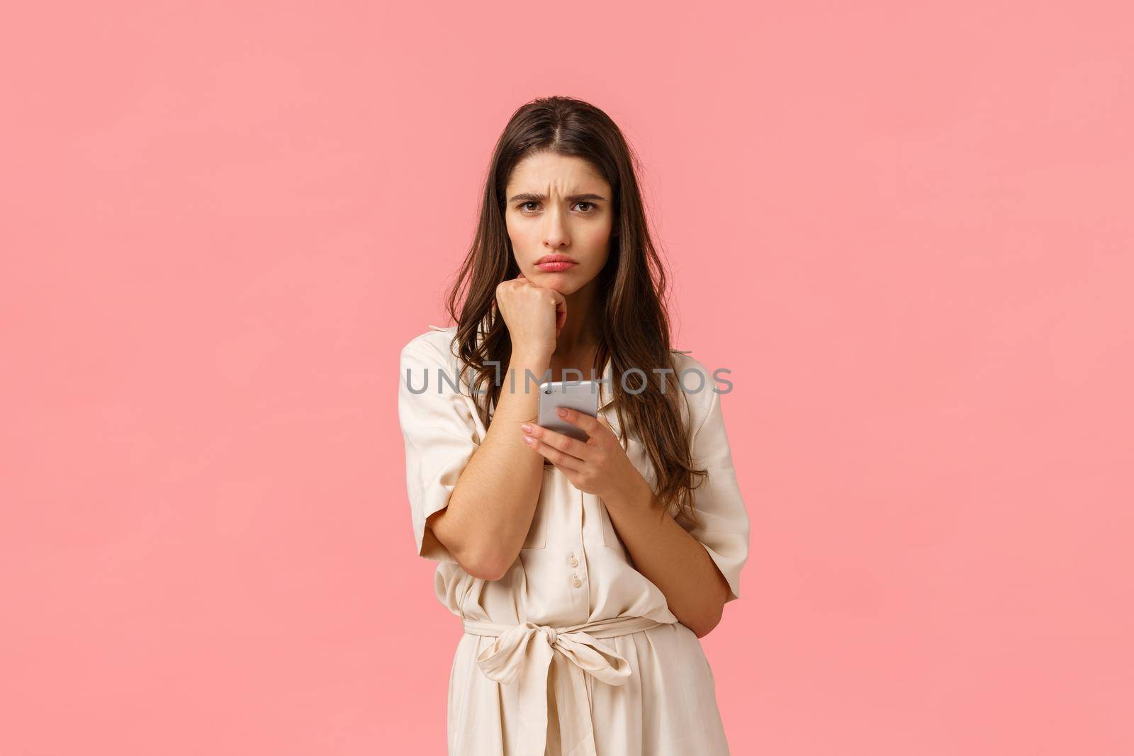 Sulky cute girlfriend facing complicated decision, frowning and sulking leaning on fist, holding smartphone, read strange message cant understand, feel perplexed, standing pink background.