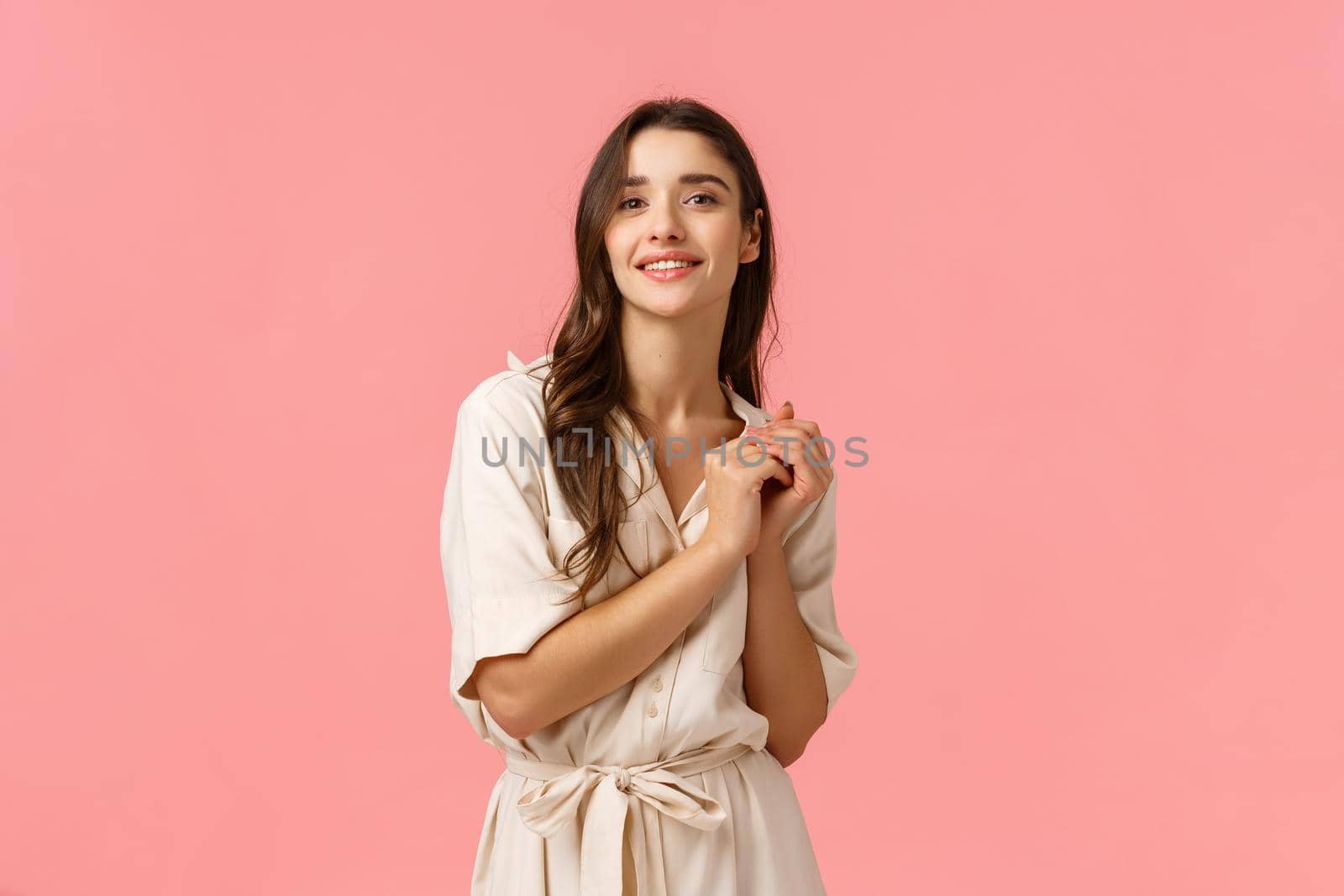 Tenderness, beauty and fashion concept. Charming tender young caucasian woman in dress, touching hands smiling sensually and gazing camera with dreamy look, standing pink background.