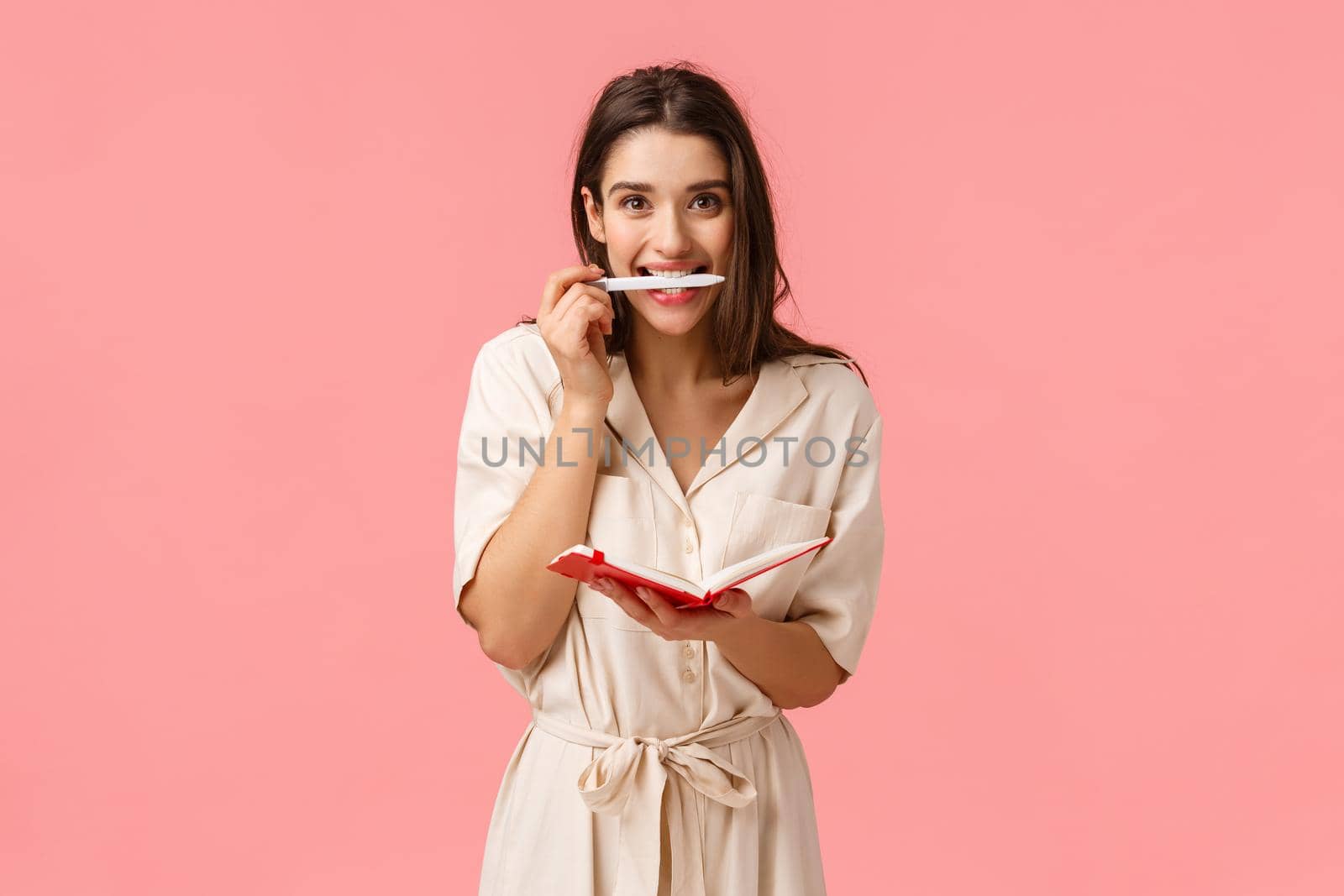 Excited and cheerful young girl stydying new language, smiling holding pen in teeth, writing in cute red notebook, standing pink background, creating personal schedule, taking notes by Benzoix