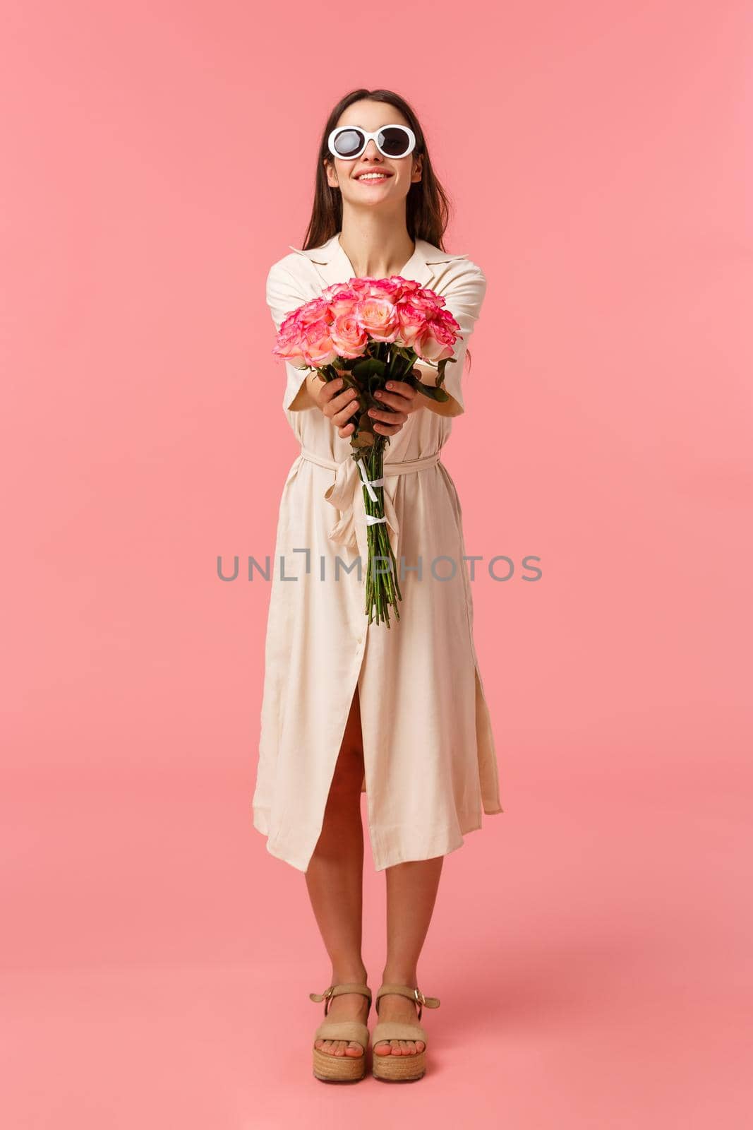 Full-length vertical portrait attractive sassy and stylish brunette woman giving flowers to her girlfriend, extending hands towards camera holding nice bouquet roses, standing pink background by Benzoix