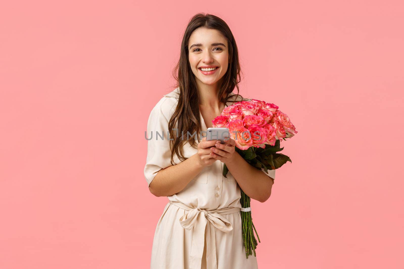 Technology, romance and happiness concept. Tender young cheerful smiling woman with beautiful flowers, holding smartphone, answer on congratulations b-day, chatting on birthday, pink background.