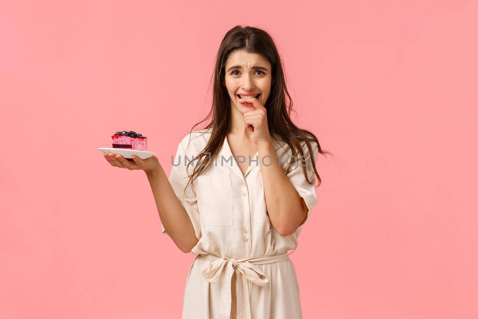Maybe just one bite. Tempting and eager brunette woman want try tasty piece cake, holding dessert frowning and biting fingernails from desire to eat sweets, resist trying stick diet, pink background.