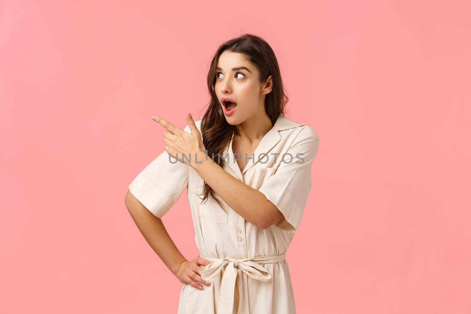 Impressed and astonished, smiling pretty female with dark curly hair, perfect skin, wear dress, pointing and looking upper left corner amused, seeing wonderful shoes matching outfit, pink background by Benzoix